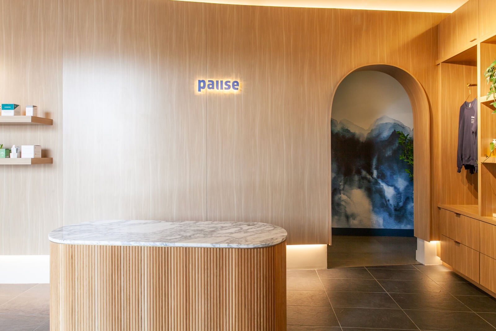 Reception area with a marble-topped counter, wood-paneled walls, illuminated "pause" sign, shelves with products, and an arched entryway leading to a mural of mountains.