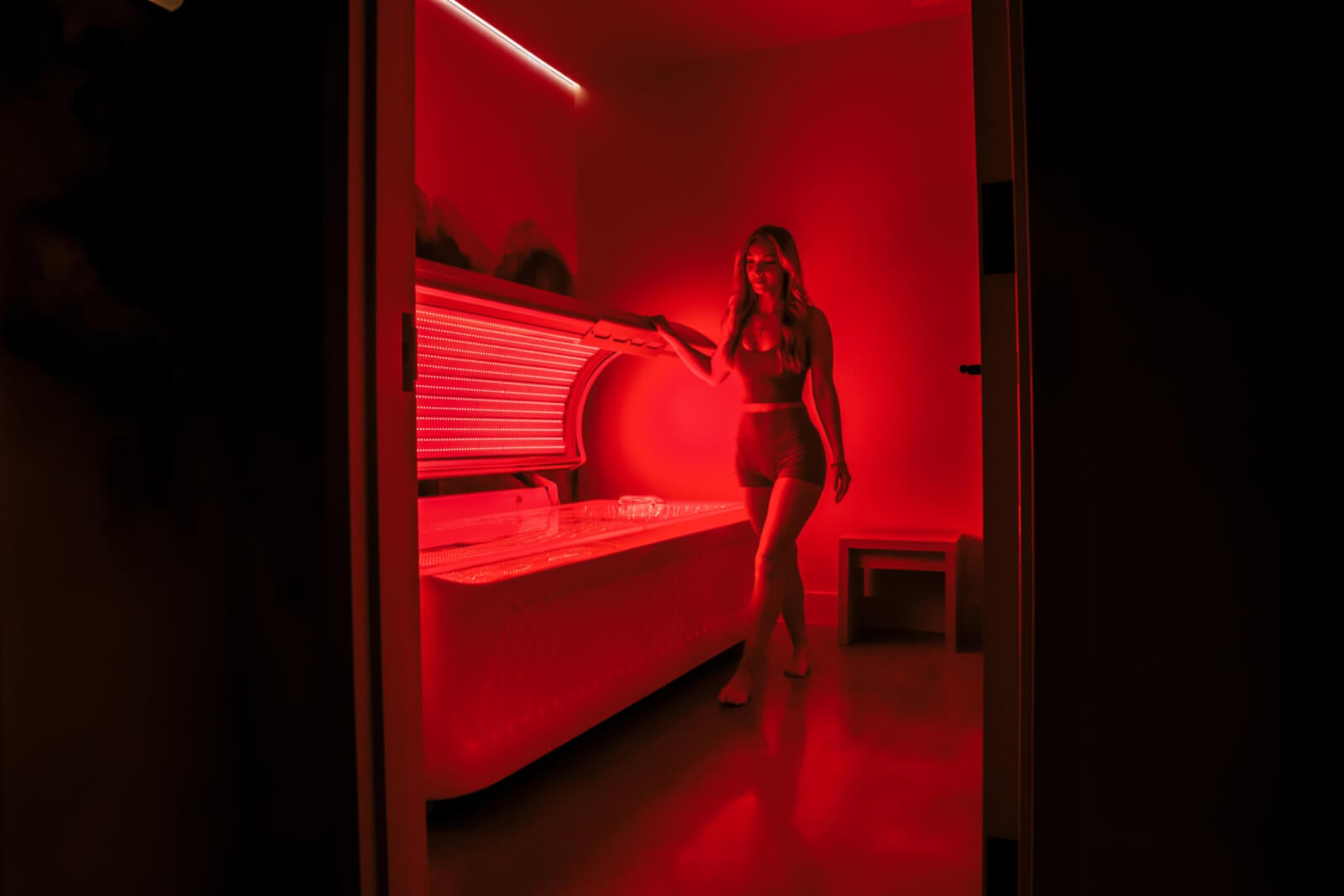 A woman in a swimsuit stands by an open tanning bed in a dimly lit room illuminated by red light.