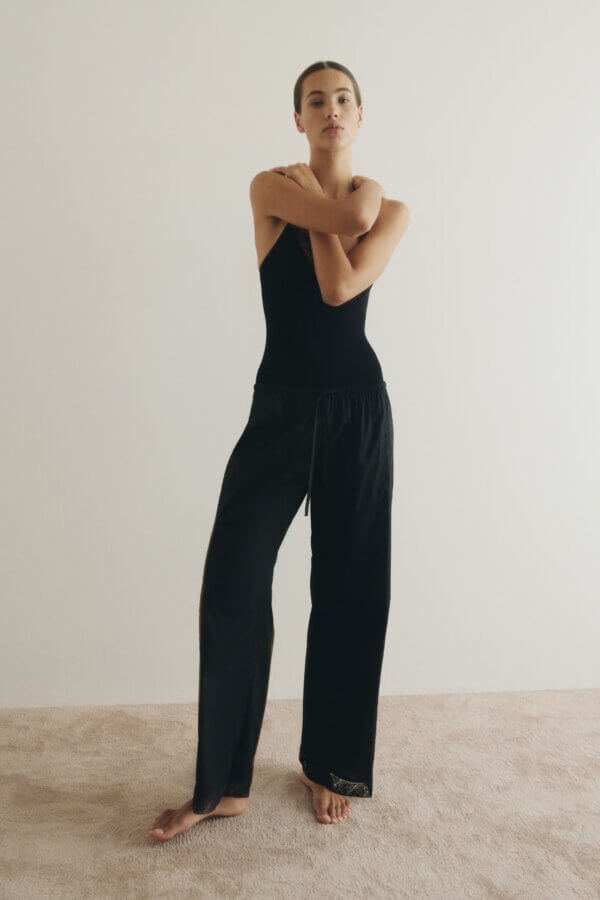 Trend Alert: Lace is Back! - 8 A person in a black sleeveless top with lace detailing and loose black pants stands barefoot on a light carpet, arms crossed over their chest, in front of a plain light-colored wall—a subtle fashion trend alert.