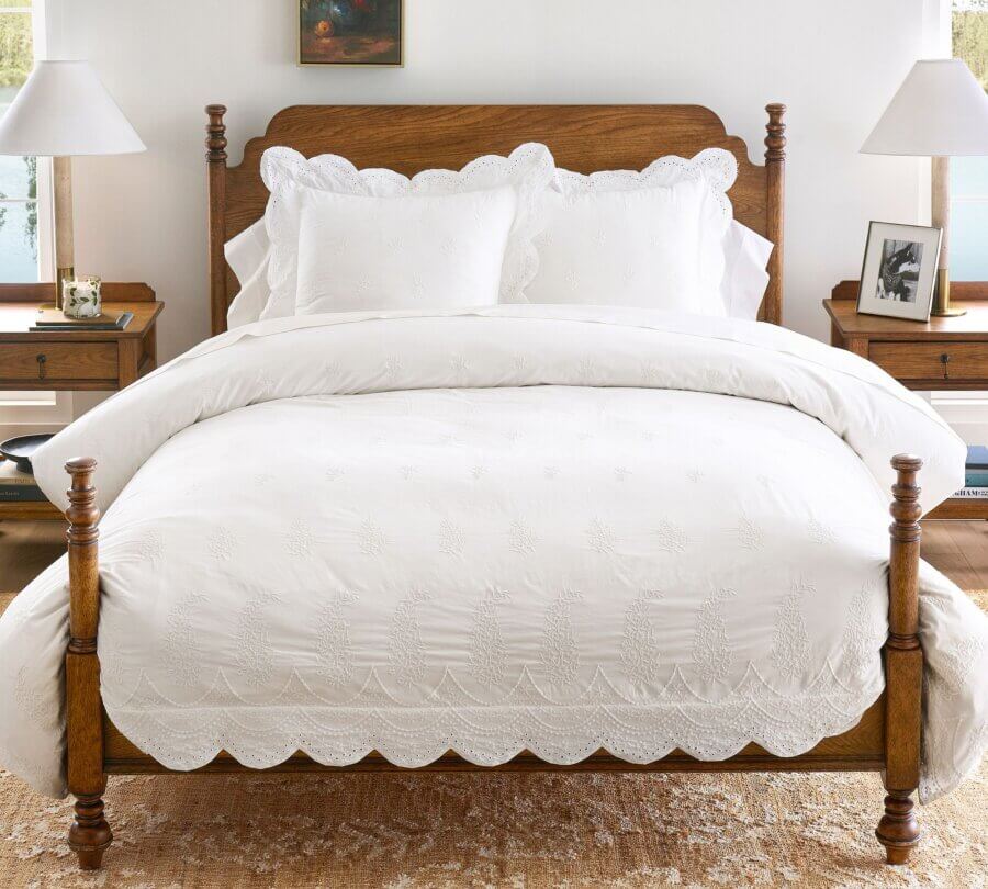 Trend Alert: Lace is Back! - 26 A wooden bed with white, scalloped-edge bedding and two matching pillows—featuring subtle lace details—is centered between two wooden nightstands in a bright bedroom.