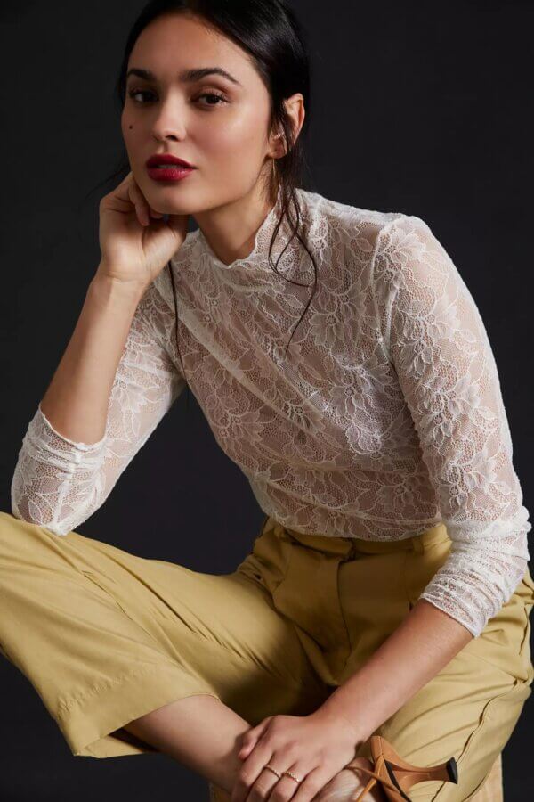 Trend Alert: Lace is Back! - 3 A woman poses against a dark background in a chic white lace long-sleeve top and beige pants, her hand thoughtfully resting on her chin.