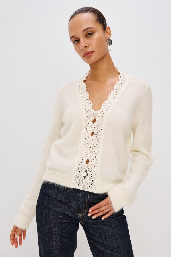 Trend Alert: Lace is Back! - 11 Trend alert: A woman showcases chic fashion in a cream-colored sweater with a scalloped lace-trimmed V-neck and dark jeans, standing against a plain light background.