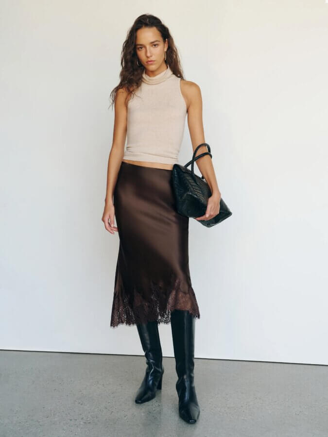 Trend Alert: Lace is Back! - 7 Lace trend: A woman stands against a white wall wearing a sleeveless beige top, a dark brown satin midi skirt with Lace trim, black knee-high boots, and holding a black handbag.