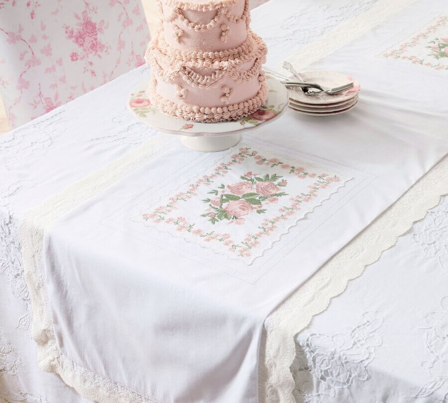 Trend Alert: Lace is Back! - 24 A pink frosted cake sits on a floral cake stand atop a trendy white lace tablecloth with a floral runner, beside a stack of plates and spoons.