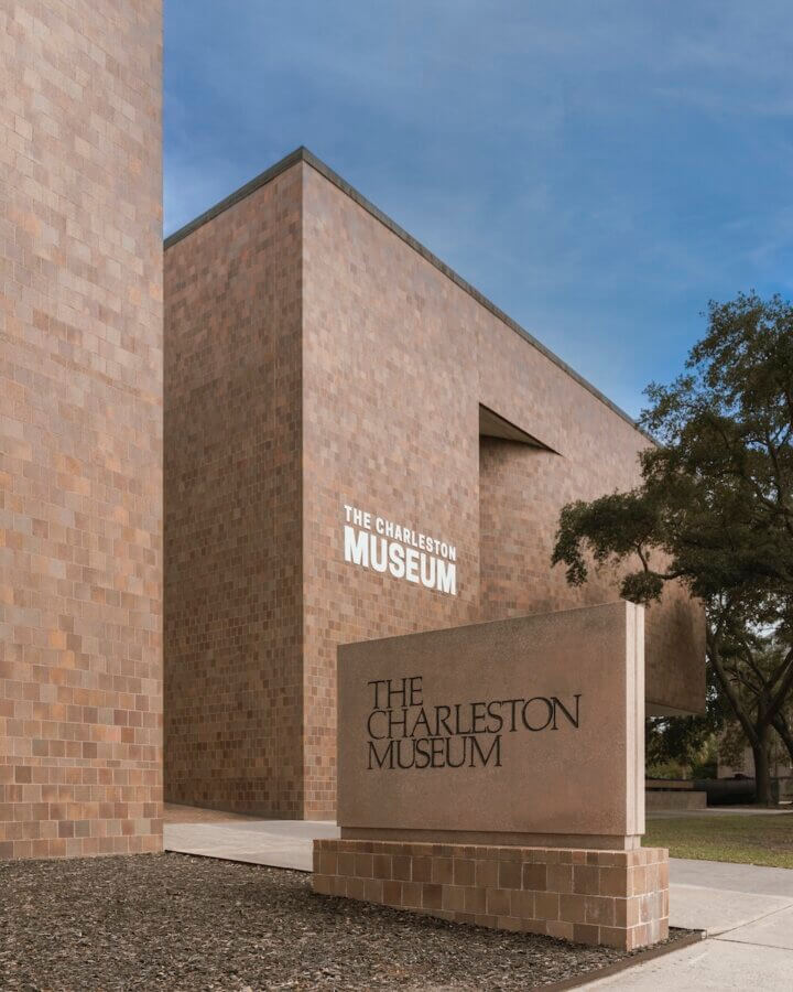 5 Must-Visit Lowcountry Attractions for Exploring Revolutionary History - 2 Modern brick building with "The Charleston Museum" sign on the exterior wall and a matching stone sign in front, surrounded by trees and a clear sky—one of the must-see Charleston attractions.