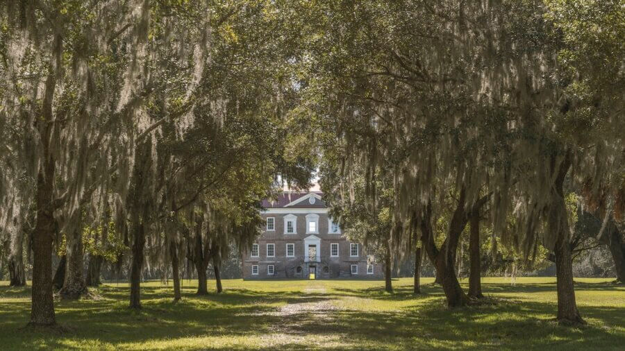 5 Must-Visit Lowcountry Attractions for Exploring Revolutionary History - 6 A brick mansion with white trim, a classic example of Charleston attractions, is seen through a path lined with large trees draped in Spanish moss on a sunny day.