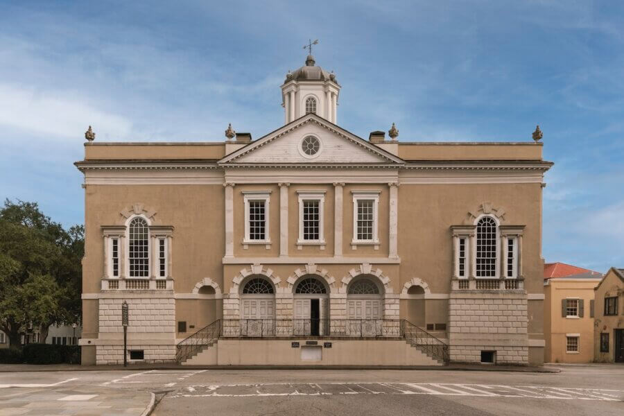 5 Must-Visit Lowcountry Attractions for Exploring Revolutionary History - 3 A symmetrical historic building with arched windows, three entry doors, a central cupola, and a grand staircase leading to the main entrance—one of the notable Charleston attractions set against a clear sky.