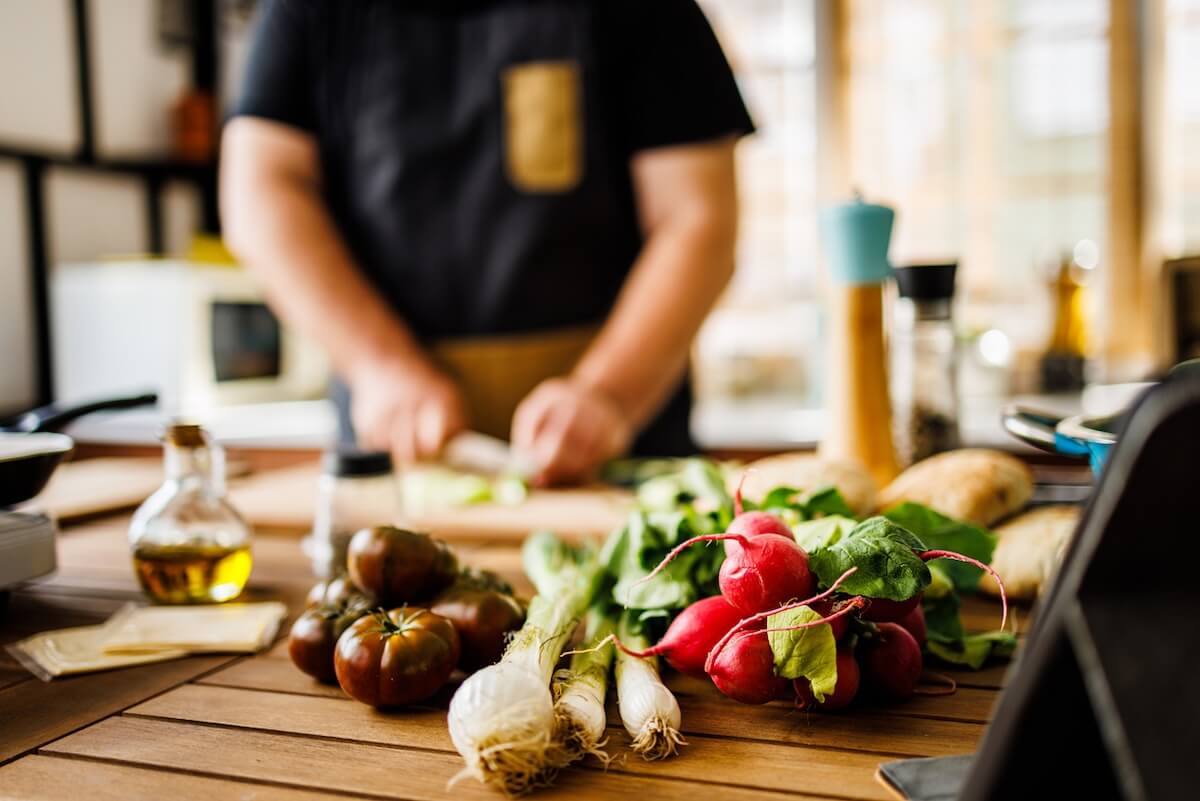 A person wearing an apron chops vegetables on a wooden kitchen counter with fresh produce and cooking ingredients in the foreground—a kitchen tip from a Veteran BHAM Chef for a cleaner kitchen.