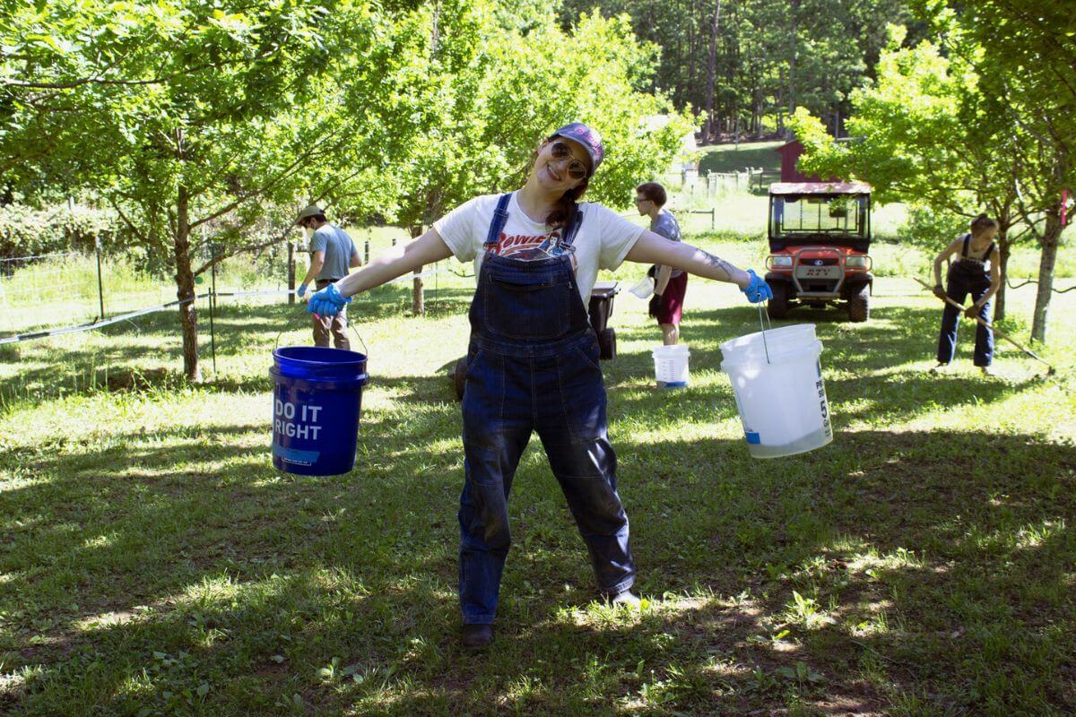 A person in overalls stands outdoors holding two buckets, arms outstretched, during truffle hunting in the South. Others work in the background among trees on a sunny day, showcasing one of the region’s unique places.