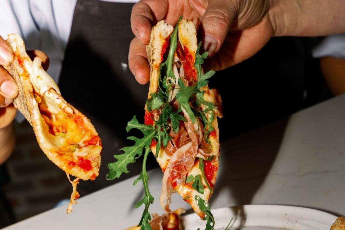 Two hands hold folded slices of pizza topped with arugula, cured meat, tomato sauce, and melted cheese over a white plate—a delicious highlight from one of the new restaurants in Birmingham.