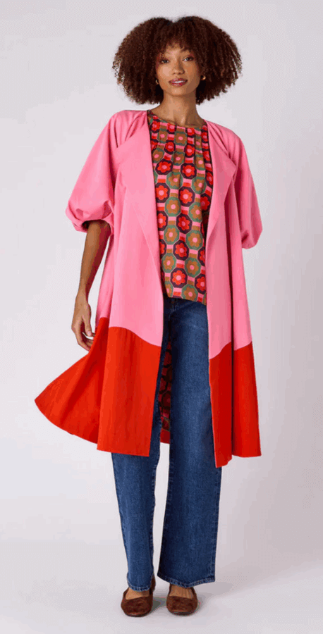 17 Birmingham FINDS for the New Year - 2 A woman wearing a long pink and red coat, a patterned blouse, blue jeans, and brown shoes stands against a plain background.