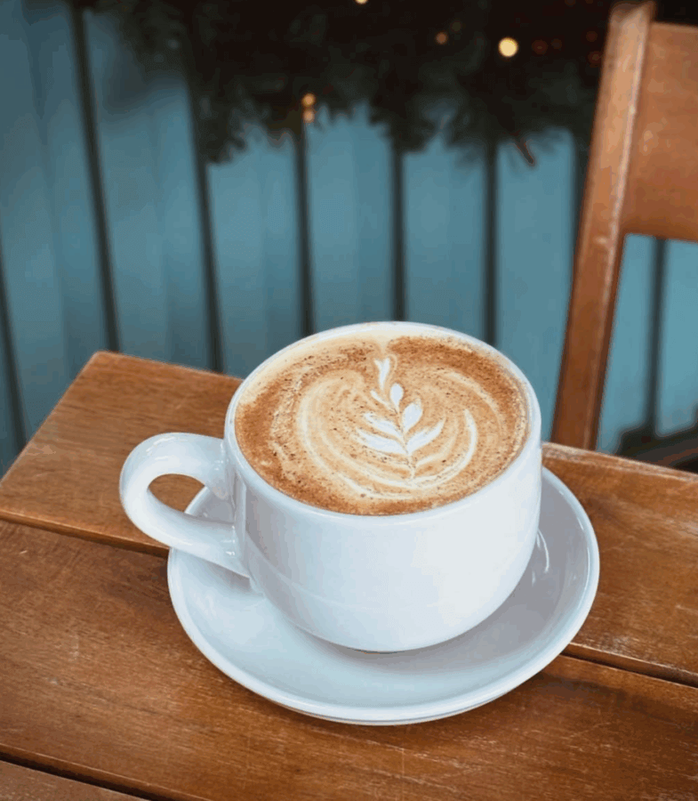 10 New Birmingham Restaurants to Try This Month - 20 A cup of coffee with leaf-shaped latte art sits on a white saucer atop a wooden table—perfect for discovering new restaurants or cozy cafes among the best Birmingham restaurants, with a wooden chair and blurred background nearby.