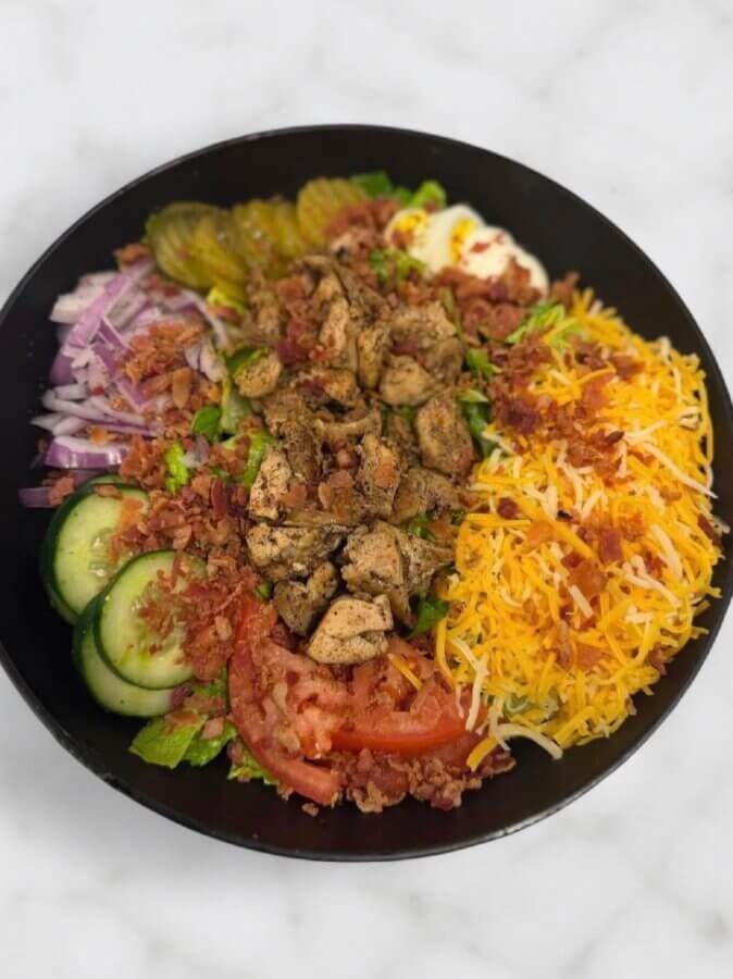 10 New Birmingham Restaurants to Try This Month - 24 A black bowl filled with chopped grilled chicken, shredded cheese, bacon bits, cucumber, tomato, red onion, pickles, and hard-boiled eggs on greens—perfect for those exploring new restaurants in Birmingham.