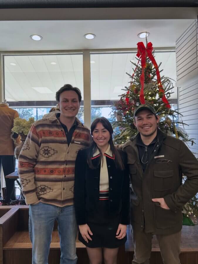 Avontuur: The New Homewood Shop You Need to Know - 3 Andrew Adams, Anna Kolbie, and Luke Kolbie stand smiling in front of a decorated Christmas tree indoors, large windows behind them, celebrating the exquisite craftsmanship found at the Homewood specialty shop.