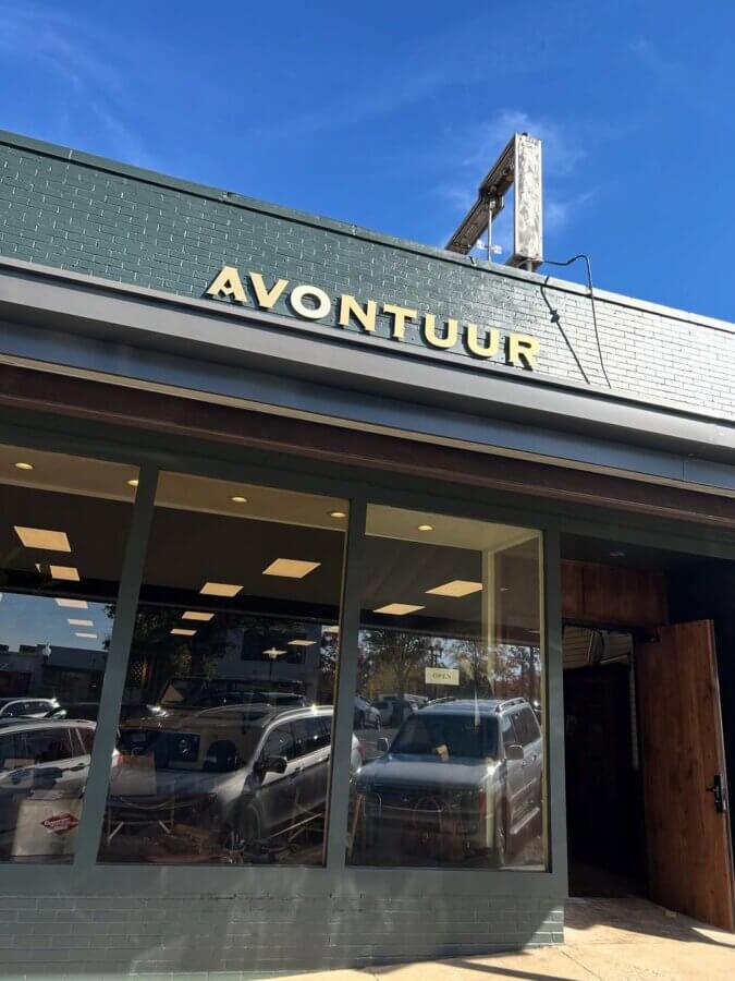 Avontuur: The New Homewood Shop You Need to Know - 2 The exterior of a building with large windows and a green sign above the entrance displaying the word "AVONTUUR" in yellow letters hints at a new shop renowned for its exquisite craftsmanship.