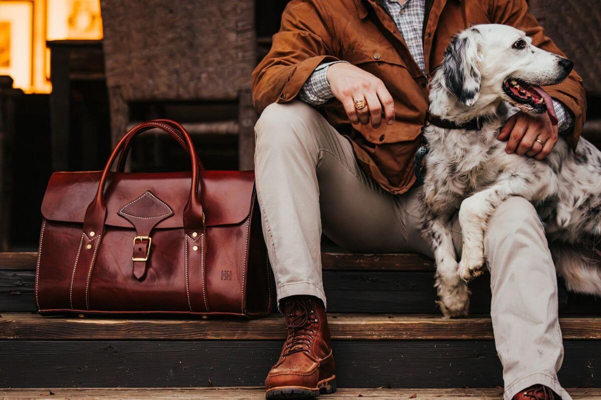 A person in a brown jacket and boots sits on stairs with a dog, holding its paw. Nearby rests a brown leather bag, products featured at Homewood's newest specialty store, Avontuur.