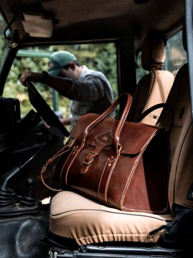 Avontuur: The New Homewood Shop You Need to Know - 4 A brown leather duffel bag with exquisite craftsmanship sits on the front passenger seat of a car, while a person in a cap adjusts something near the driver’s seat, highlighting quality found at a Homewood specialty shop.