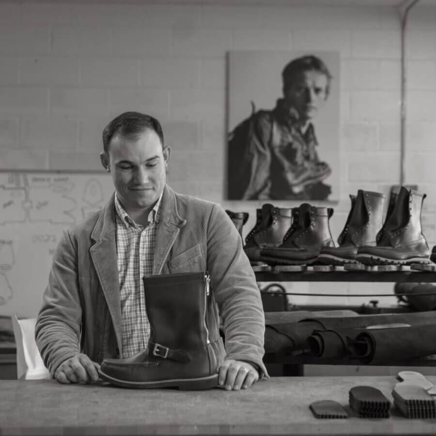 Avontuur: The New Homewood Shop You Need to Know - 7 Luke Kolbie in a workshop examines a leather boot on a table, surrounded by more boots, tools, and a large black-and-white portrait.