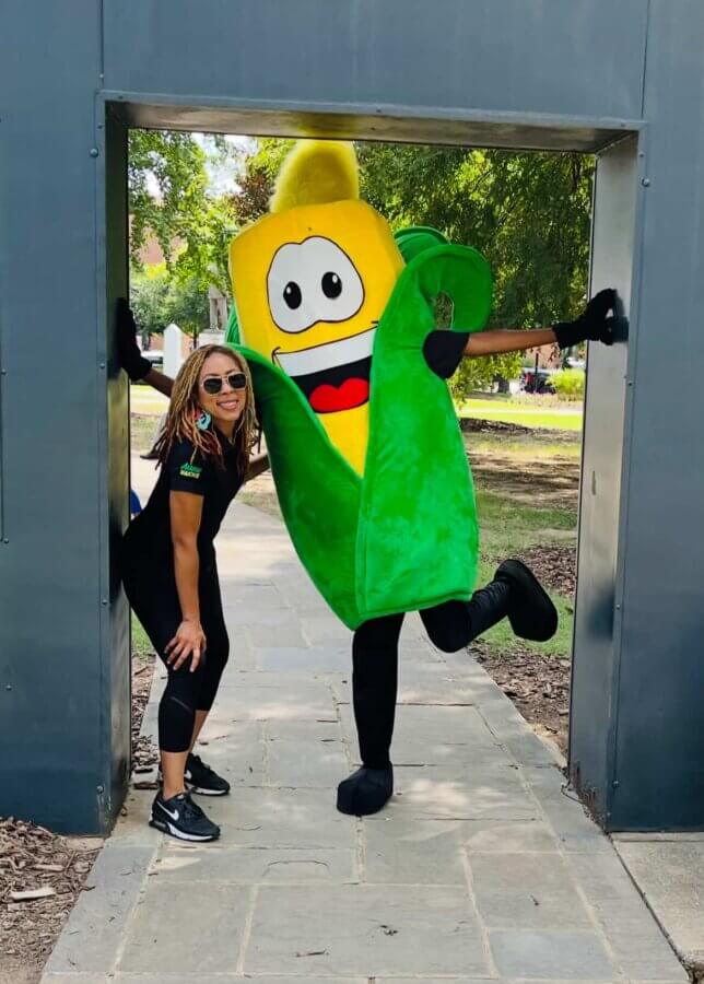 Aww Shucks! Meet the Woman Behind This Plant-Based BHAM Favorite - 2 Avrie Powell poses smiling next to a person in a green and yellow corn costume with a cartoon face, standing outdoors under a gray structure.