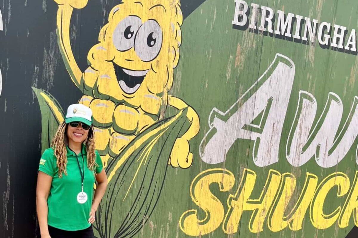 Avrie Powell in a green shirt and hat stands before a mural with a cartoon corn character and the words "Birmingham Aw Shucks," capturing the spirit of Southerners who love to eat out, especially at spots like a plant-based food truck.