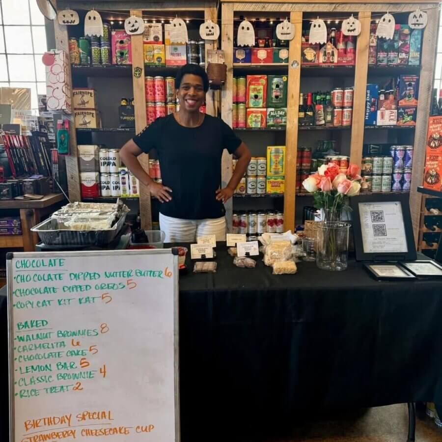 4 Decadent BHAM Chocolate Shops You Need to Know About - 3 A woman stands behind a table displaying baked goods and a menu inside a store with shelves of canned products.