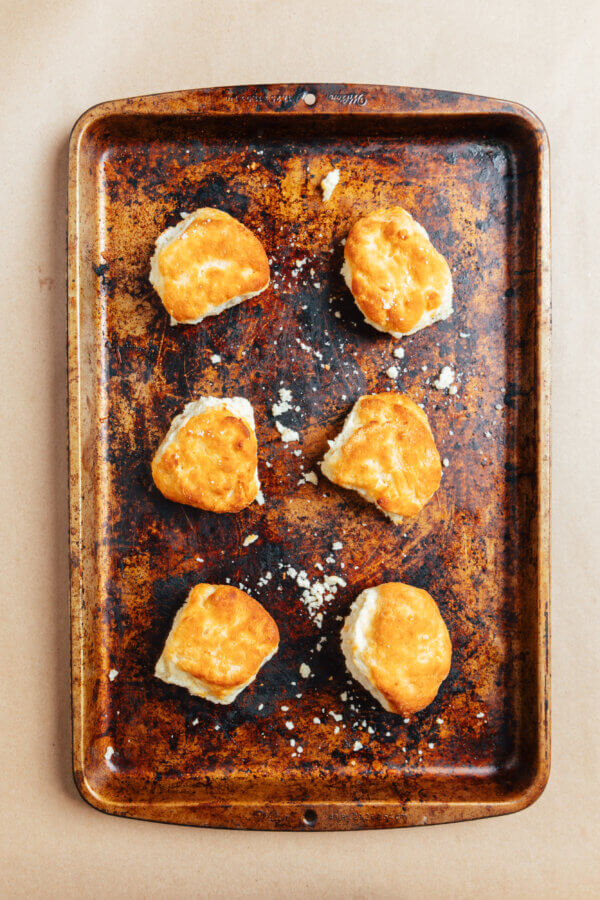 A Southern Chef's Secret to a Cleaner Kitchen in 2026 (It’s Not What You Think) - 8 Six golden-brown biscuits arranged on a worn, rectangular baking sheet with scattered crumbs, set on a light brown surface—a top tip from a Veteran BHAM Chef for a cleaner kitchen: line your pan for easy cleanup.