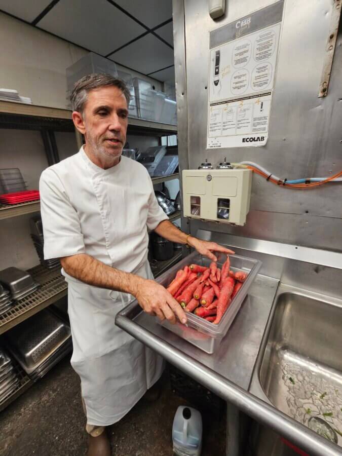 A Southern Chef's Secret to a Cleaner Kitchen in 2026 (It’s Not What You Think) - 6 Chef Chris Melville stands in a kitchen, holding a bin filled with red carrots next to a stainless steel sink.