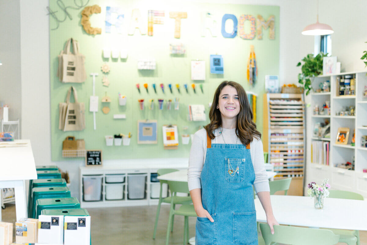 Blakely O'Connor wearing a denim apron stands in The Craft Room surrounded by art supplies and storage boxes—an inspiring space where this BHAM Women crafting enthusiast brings creative visions to life.