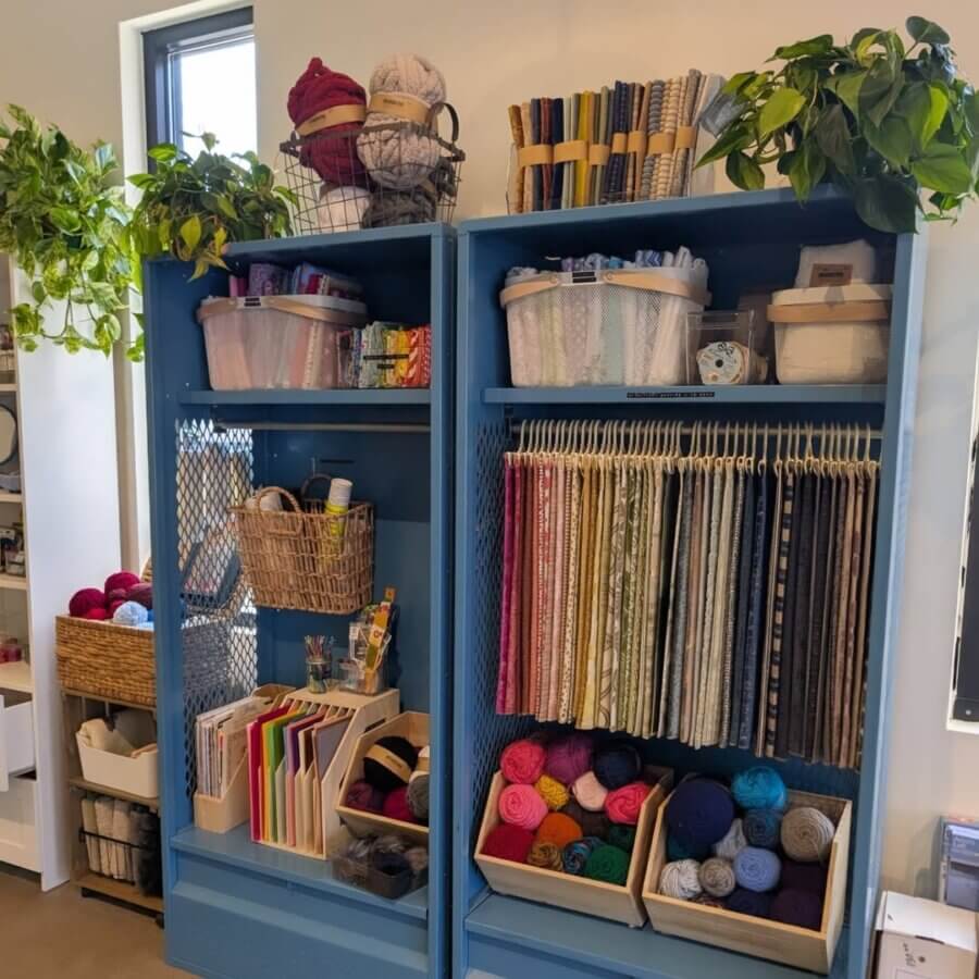 This BHAM Engineer Creates Community Through ... Crafting! - 4 Two blue shelves showcase rolled fabrics, yarn, baskets with crafting supplies, and plants on top at The Craft Room.