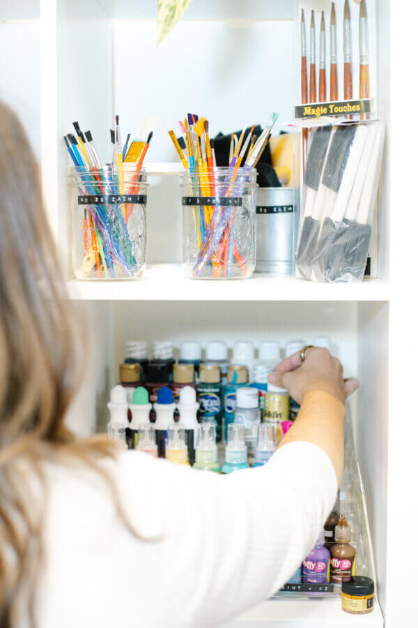 This BHAM Engineer Creates Community Through ... Crafting! - 5 A person reaches for art supplies on a shelf, with jars of paintbrushes, bottles of paint, and paint spatulas organized on different levels—perfect for crafting sessions inspired by BHAM Women.
