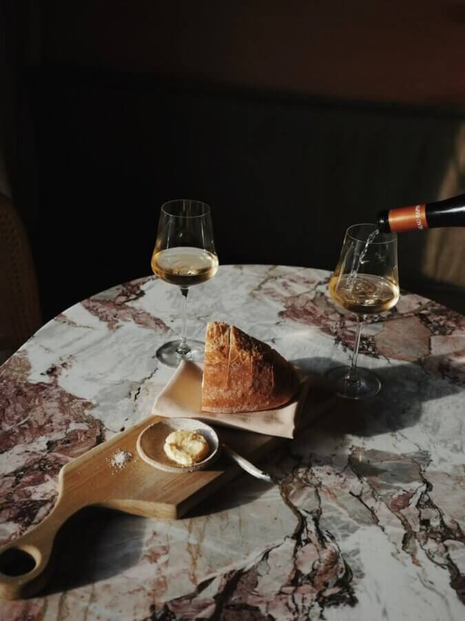 Two glasses of white wine, a piece of bread on a napkin, and a small wooden board with butter and salt are arranged on a marble table, with wine being poured into one glass.