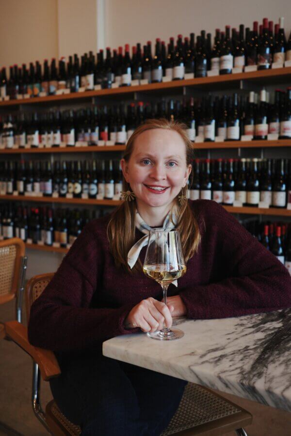 Kady Wolhfarth sits at a marble table holding a glass of white wine, with shelves of wine bottles in the background.