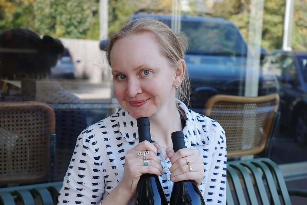 A woman with light hair and blue eyes holds two bottles of golden age wine while sitting outdoors at a table with cane chairs and parked cars visible in the background.