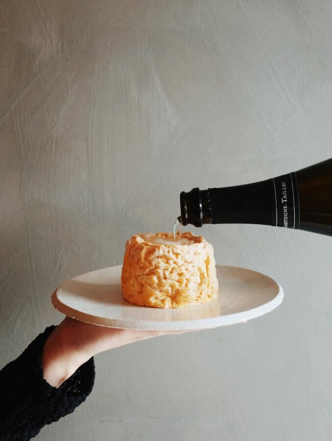 A hand holds a plate with a round cheese while sparkling wine is being poured onto it from a bottle against a plain background.