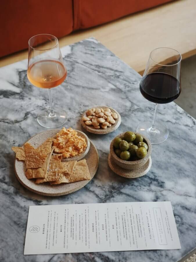 Two glasses of wine, one rosé and one red, with plates of crackers and cheese, green olives, and almonds on a marble table next to a menu.