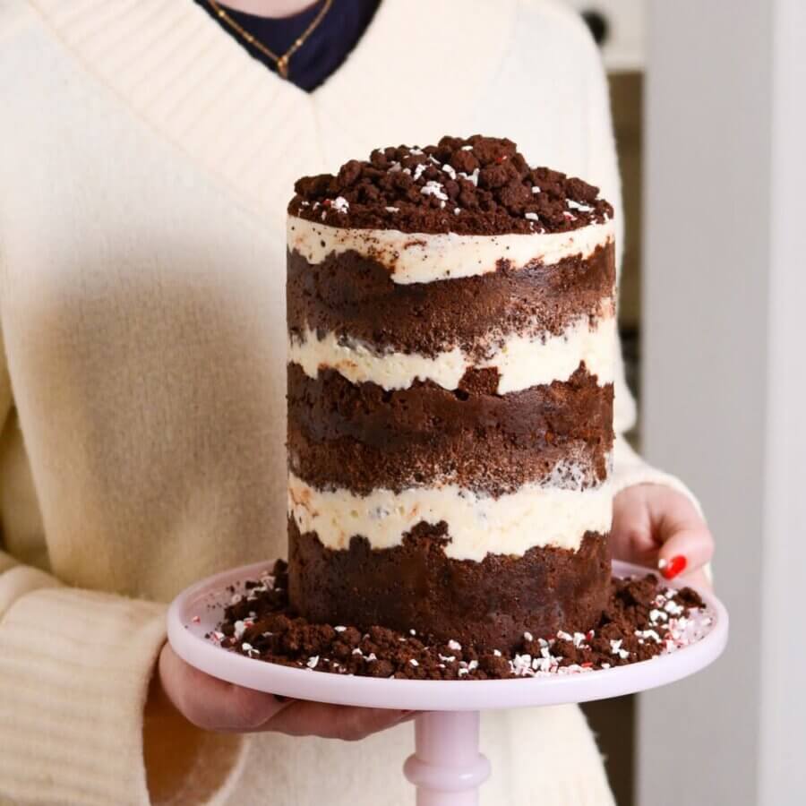 She Bakes Some of BHAM’s Most Iconic Wedding Cakes - 7 A person in a cream sweater holds an iconic, unfrosted chocolate layer cake with cream filling and chocolate crumbs on top, displayed on a pink cake stand.