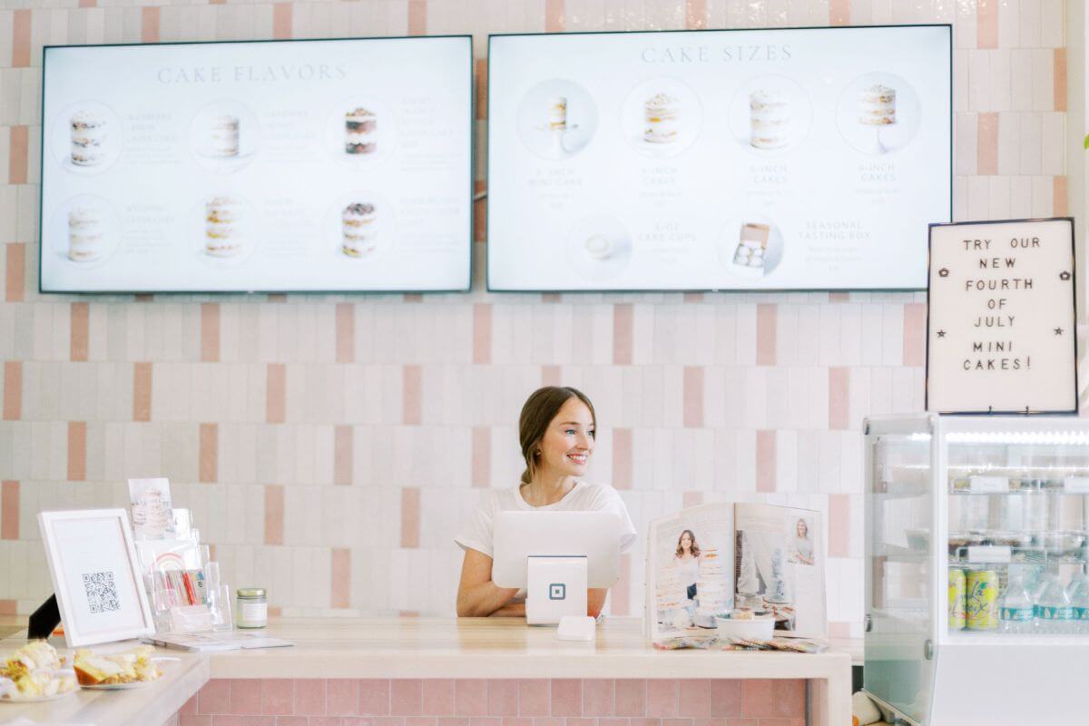Owner Mallory Mason stands behind the counter at Daughters Baking, with cake flavor and size menus displayed on screens behind her and a sign promoting new mini cakes, highlighting their iconic wedding cakes.