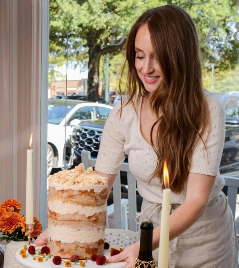 She Bakes Some of BHAM’s Most Iconic Wedding Cakes - 2 Mallory Mason in a white top arranges an iconic, tall layered wedding cake decorated with flowers and crumbles on a table set with candles and a bottle, near a window overlooking trees and parked cars.