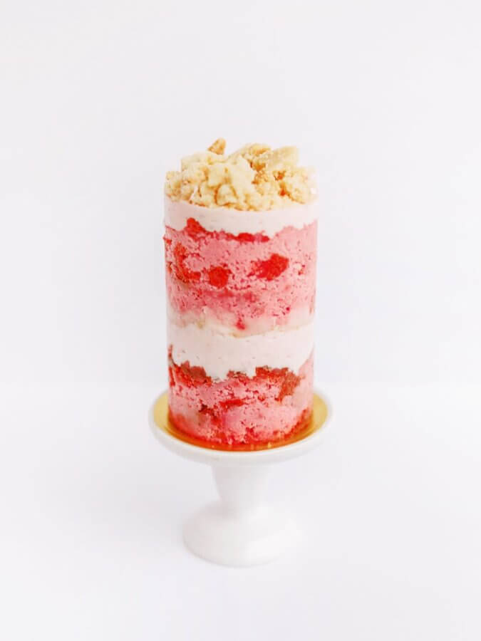 She Bakes Some of BHAM’s Most Iconic Wedding Cakes - 5 A tall, cylindrical layered cake with iconic pink and white layers, topped with crumbled pieces, is elegantly displayed on a small white pedestal stand.