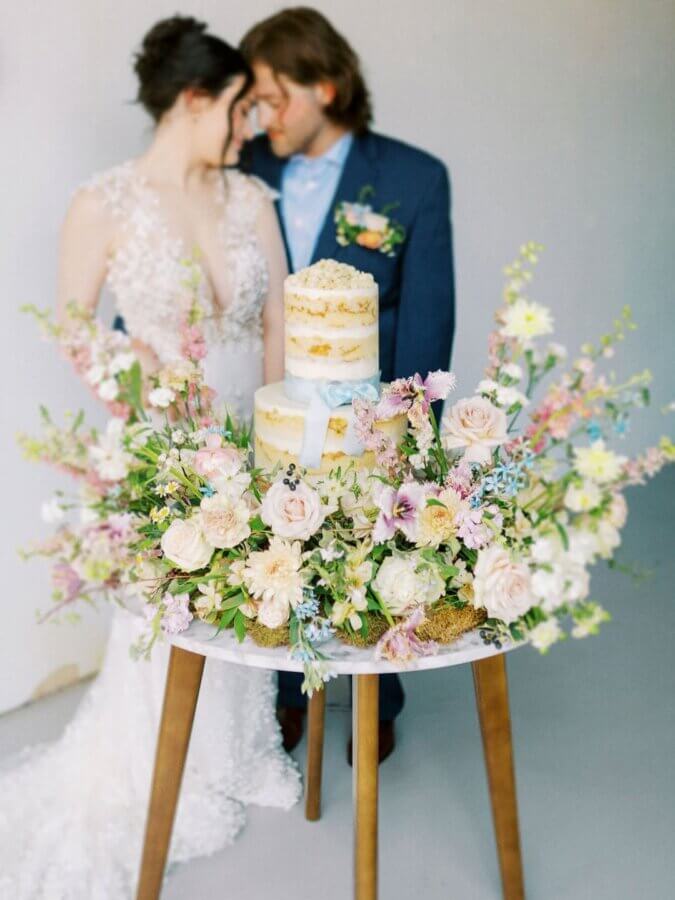 She Bakes Some of BHAM’s Most Iconic Wedding Cakes - 3 The bride and groom stand closely behind a small table in BHAM, beautifully decorated with flowers and an iconic two-tiered wedding cake.