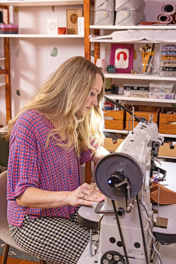 Cold Gold Standard: Bold Leather Bags & Jewelry - 5 A woman with long blonde hair operates a sewing machine in a well-organized craft room filled with bold leather bags and storage boxes, while shelves showcase her handmade jewels in the background.