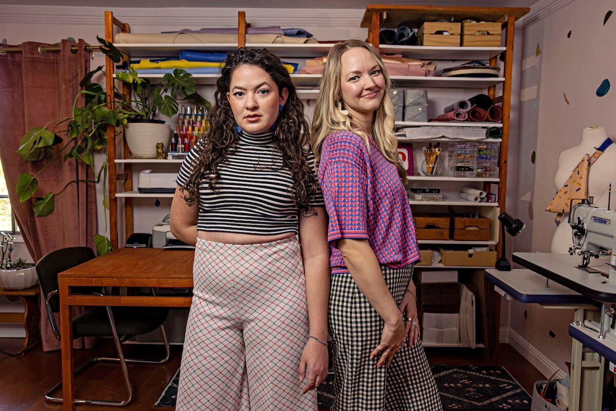 Two women stand back-to-back in a sewing studio, surrounded by shelves with fabrics, tools, leather bags, a sewing machine, and a large potted plant.