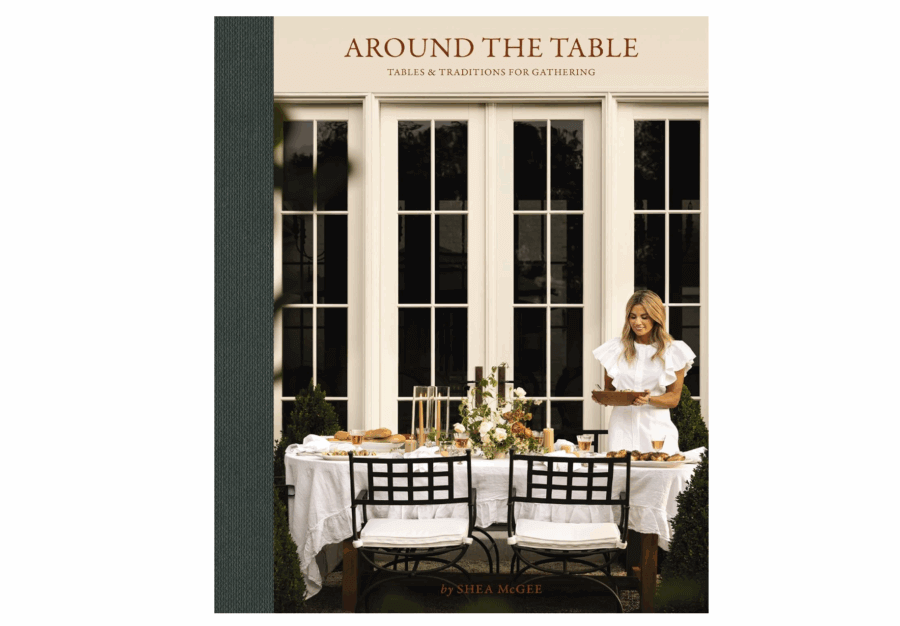 2026 Cookbook Releases We Can't Wait to Snag - 11 A woman in a white dress sets a dining table outdoors in front of tall windows, featured on the cover of "Around the Table" by Shea McGee—one of the most anticipated new cookbooks among 2026 cookbook releases.
