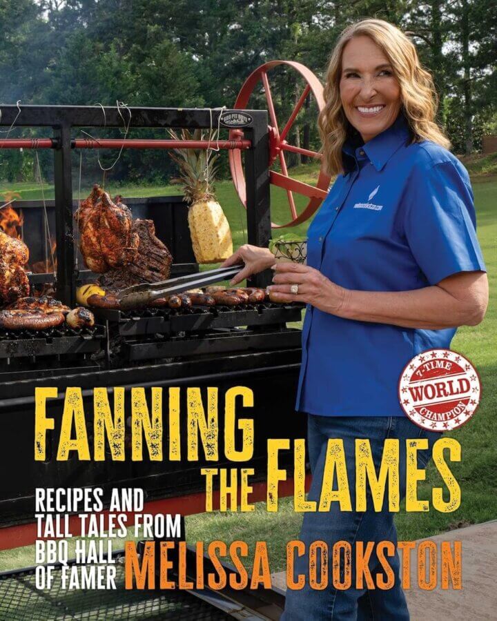 2026 Cookbook Releases We Can't Wait to Snag - 12 A woman in a blue shirt grills assorted meats outdoors, holding tongs and smiling. The text reads, "Fanning the Flames—Recipes and Tall Tales from BBQ Hall of Famer Melissa Cookston," one of the must-have 2026 cookbooks.