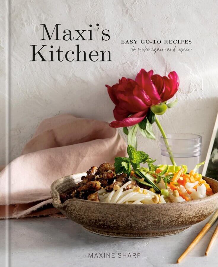 2026 Cookbook Releases We Can't Wait to Snag - 4 A cookbook titled "Maxi's Kitchen" by Maxine Sharf, featured among new cookbooks, with a bowl of food, a pink napkin, a red flower, and a glass of water on a light-colored surface.