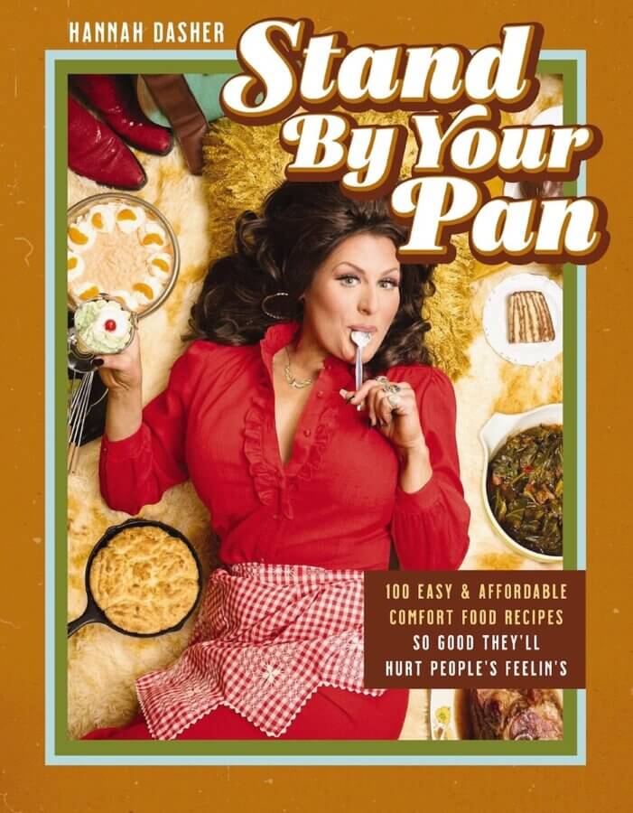 2026 Cookbook Releases We Can't Wait to Snag - 7 A woman in a red outfit with a checkered apron sits on the floor surrounded by comfort food, holding a pie and licking a spoon. The book title reads "Stand By Your Pan," one of the most anticipated upcoming cookbooks of the year.