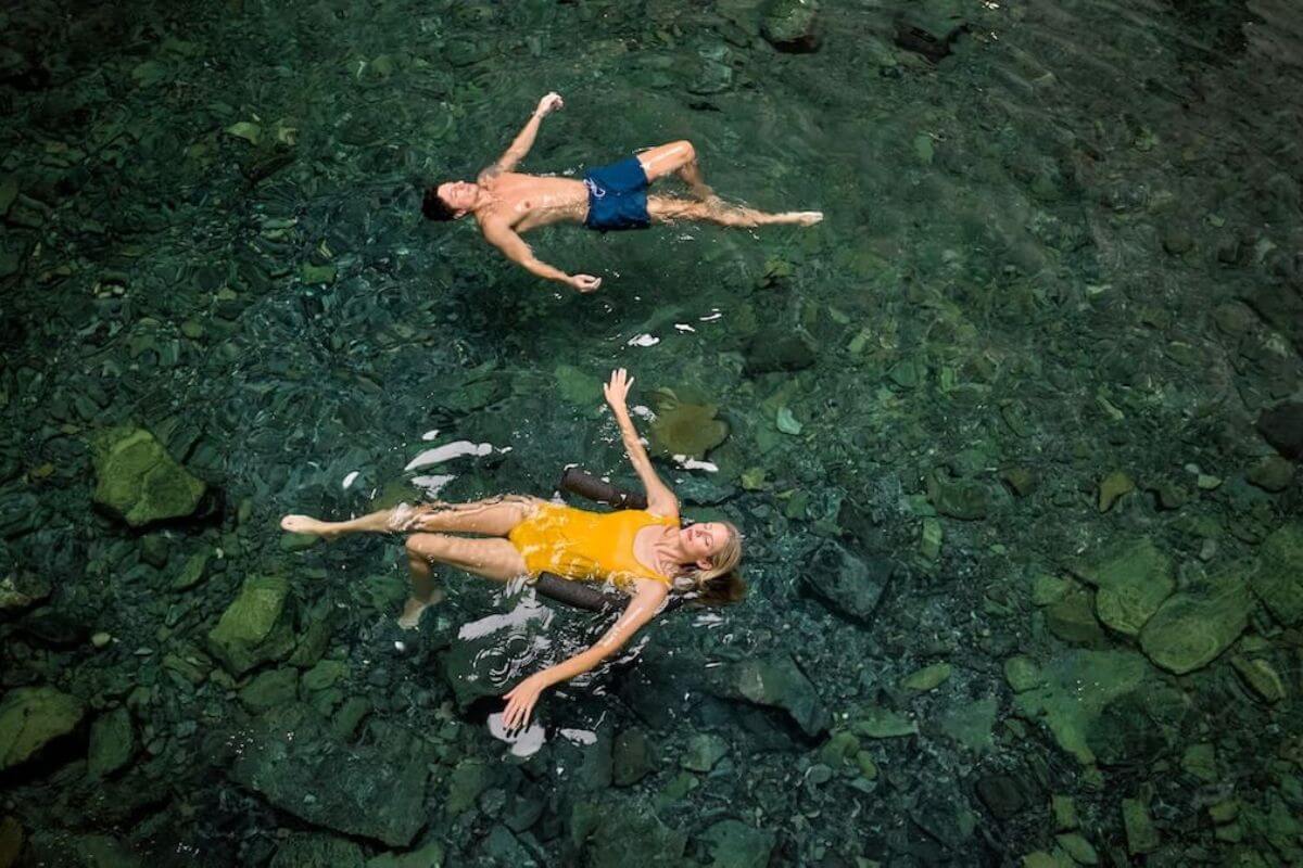 Two people float on their backs in clear water above a rocky bottom; one wears blue swim trunks, and the other a yellow swimsuit—an idyllic scene perfect for a couples getaway.
