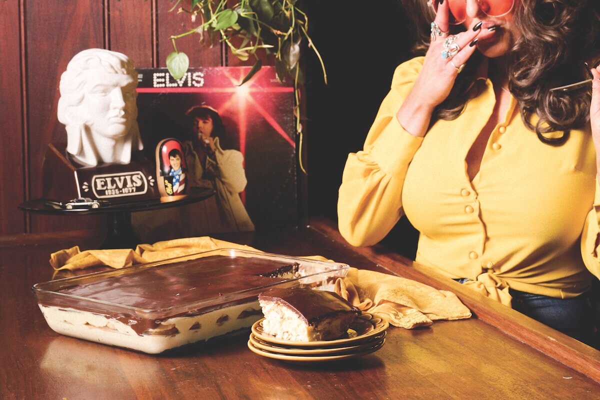 A woman in a yellow top sits at a table enjoying a chocolate dessert, surrounded by Elvis Presley memorabilia and an Elvis Éclair Cake recipe display, with an Elvis record showcased in the background.