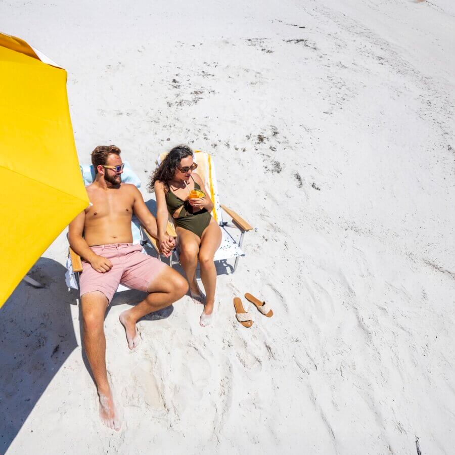 A man and a woman sit on beach chairs under a yellow umbrella, holding hands and relaxing on a sandy beach—perfect for those dreaming of fabulous beach vacations. Sandals rest nearby in the sun-warmed sand.