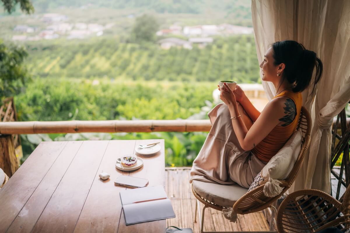 A woman sits on a terrace with coffee, reflecting on science-backed habits for life improvement. A table in front of her holds a dessert, notebook, pen, and plate as she plans her daily habits amid the green landscape.
